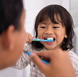 A girl child brushing her teeth looking at an adult doing the same